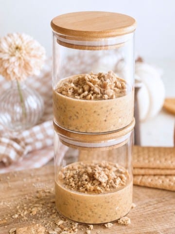Two glass jars stacked on top of each other, each filled with a creamy pumpkin overnight oats dessert topped with oat crumble. Wooden lids are on both jars, and the background includes flowers, cookies, and oats on a wooden surface.
