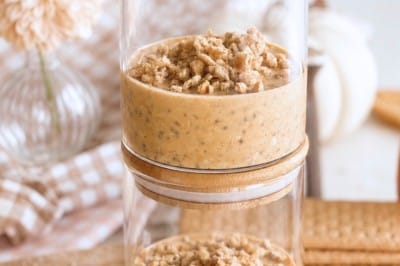Two glass jars stacked on top of each other, each filled with a creamy pumpkin overnight oats dessert topped with oat crumble. Wooden lids are on both jars, and the background includes flowers, cookies, and oats on a wooden surface.