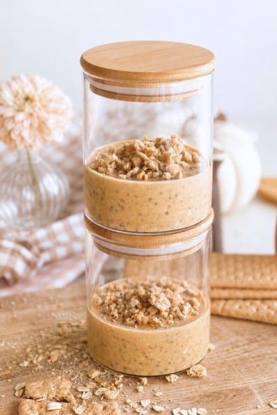 Two glass jars stacked on top of each other, each filled with a creamy pumpkin overnight oats dessert topped with oat crumble. Wooden lids are on both jars, and the background includes flowers, cookies, and oats on a wooden surface.