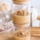 Two glass jars stacked on top of each other, each filled with a creamy pumpkin overnight oats dessert topped with oat crumble. Wooden lids are on both jars, and the background includes flowers, cookies, and oats on a wooden surface.