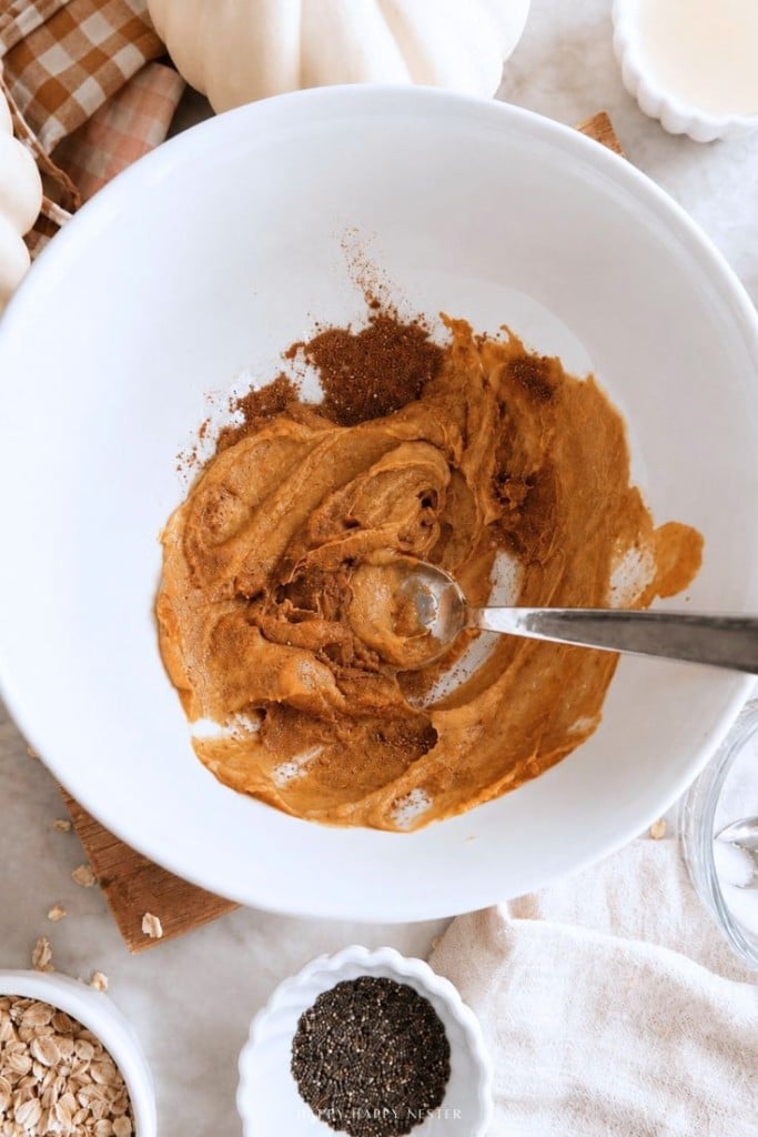 A white bowl contains a mixture of pumpkin puree and spices being stirred with a spoon, surrounded by small bowls of oats, chia seeds, and a checkered napkin on a light surface.