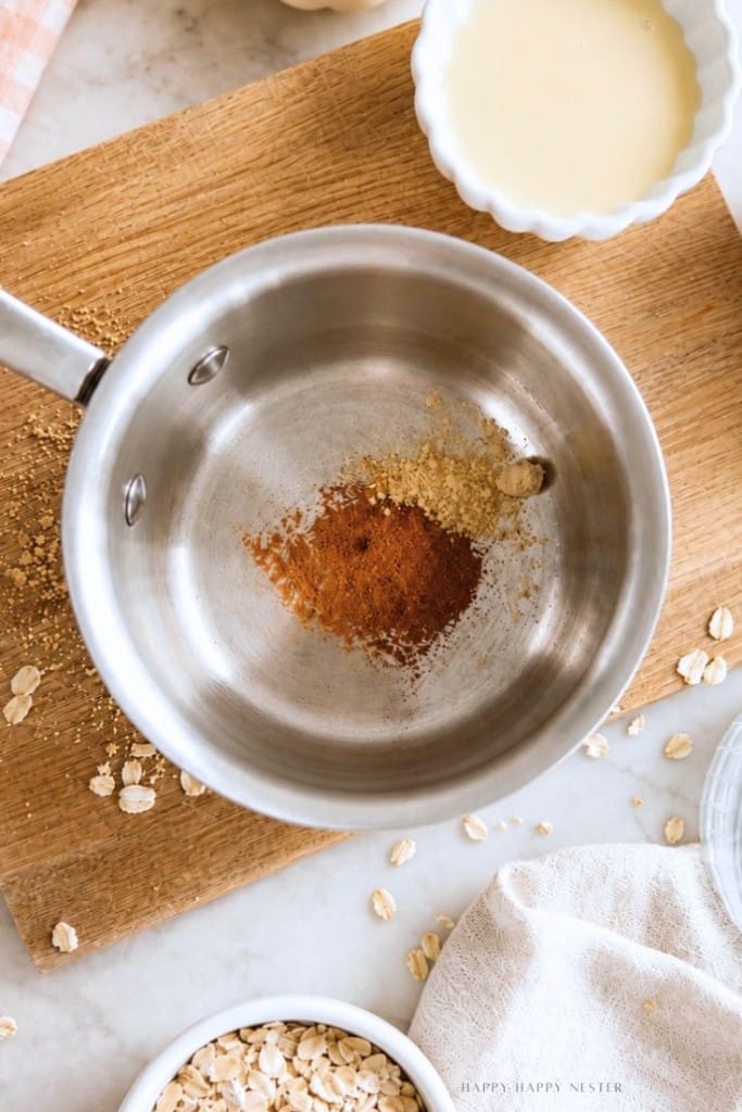 A metal saucepan with ground spices sits on a wooden board. Nearby are a bowl of oats, a bowl of liquid, some scattered oats, and a beige towel.