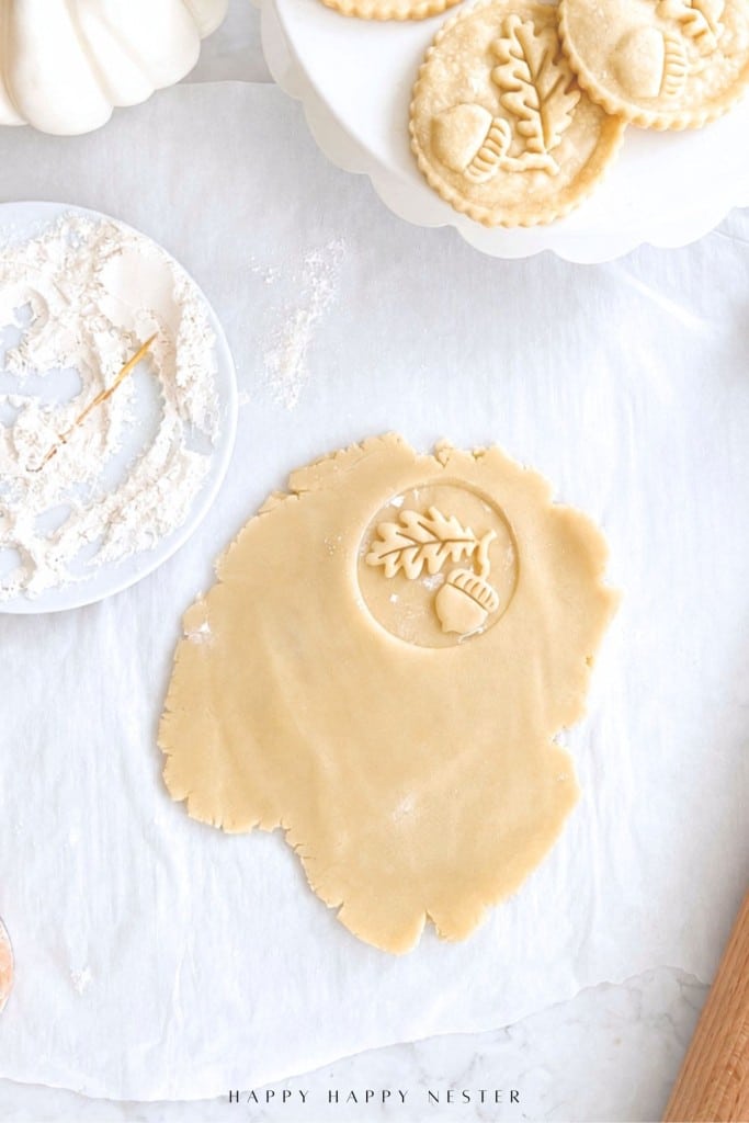 Unbaked pie dough on parchment with an embossed acorn and leaf design, surrounded by floured tools and finished decorative pie crust tops on a white plate.