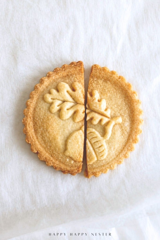 A round cookie with an embossed oak leaf and acorn design, broken neatly in half, sits on a white cloth background.