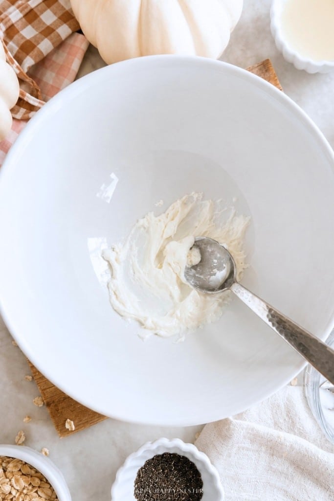 A metal spoon rests in a white bowl with a small amount of cream cheese being mixed. Surrounding the bowl are ingredients like oats, chia seeds, milk, a napkin, and a white pumpkin.