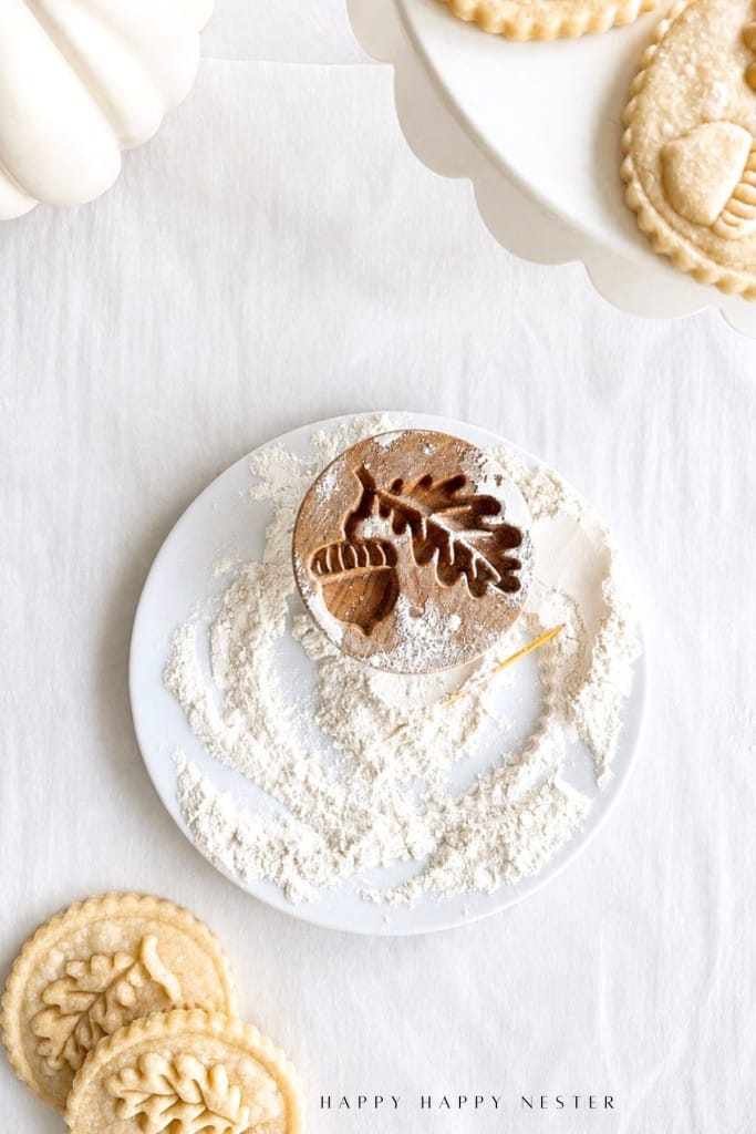 A wooden cookie stamp with an acorn and leaf design sits on a floured plate, surrounded by flour and stamped shortbread cookies with delicate leaf patterns on a white surface.