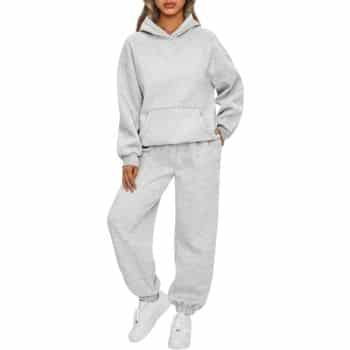 A person wearing a light gray hooded sweatshirt and matching sweatpants with white sneakers stands against a plain white background.
