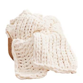 Soft ivory chunky knit throw blanket draped from a woven basket in a bright neutral room.
