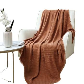 Rust-colored textured knit throw blanket draped over a white armchair in a bright living room.
