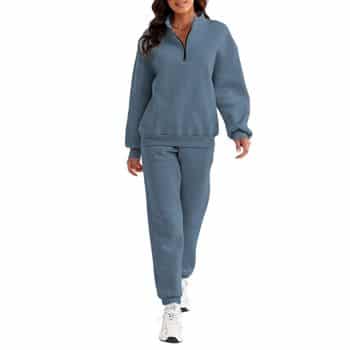 A woman wears a blue zip-up sweatshirt and matching sweatpants with white sneakers, walking forward against a white background.