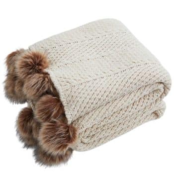 Cream knit throw blanket folded with large brown faux fur pom poms along the edge.
