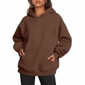 A person wearing a loose-fitting brown hoodie with front pockets and black pants, standing with hands in the pockets against a plain background.