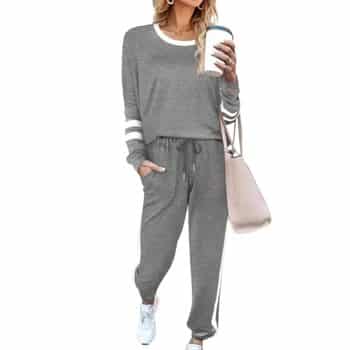 Woman wearing a gray lounge set with white stripes, holding a coffee cup and a pink shoulder bag, and wearing white sneakers. Her face is not visible.