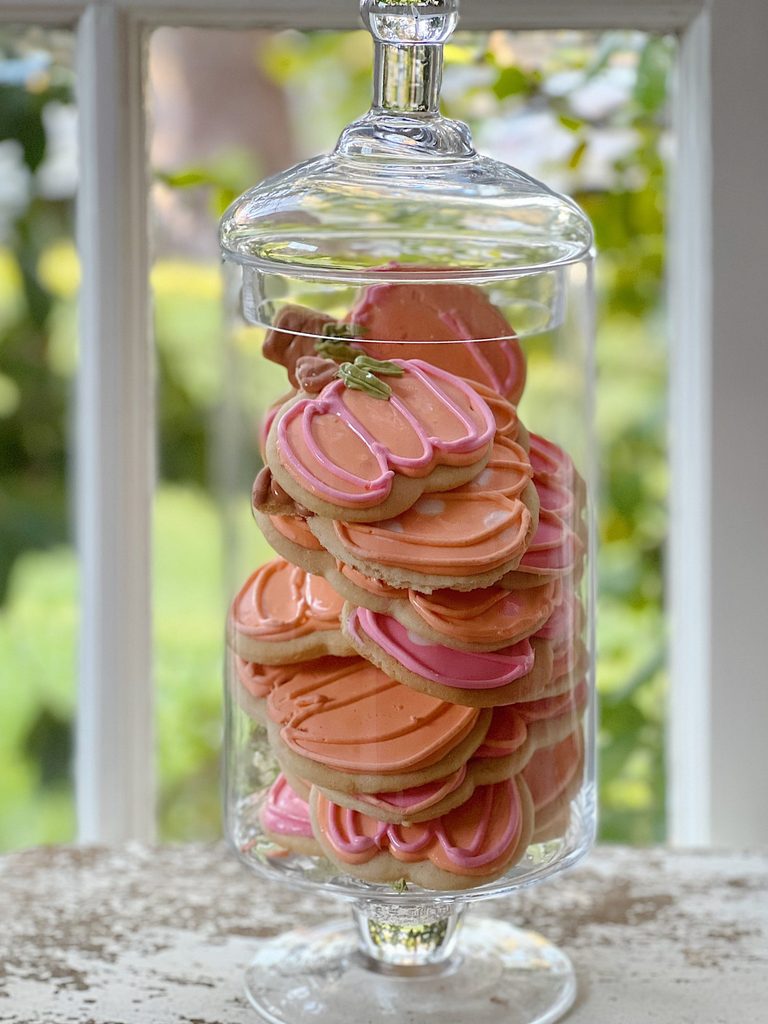 A glass jar filled with pumpkin-shaped cookies decorated with orange and pink icing, placed on a windowsill with greenery visible outside, offers charming halloween food inspiration.