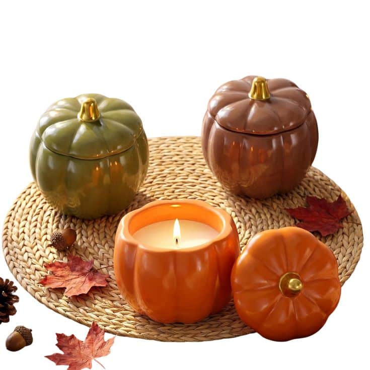 Three pumpkin-shaped containers sit on a woven mat with autumn leaves. The orange one is open, revealing a lit candle inside, while the green and brown containers are closed.