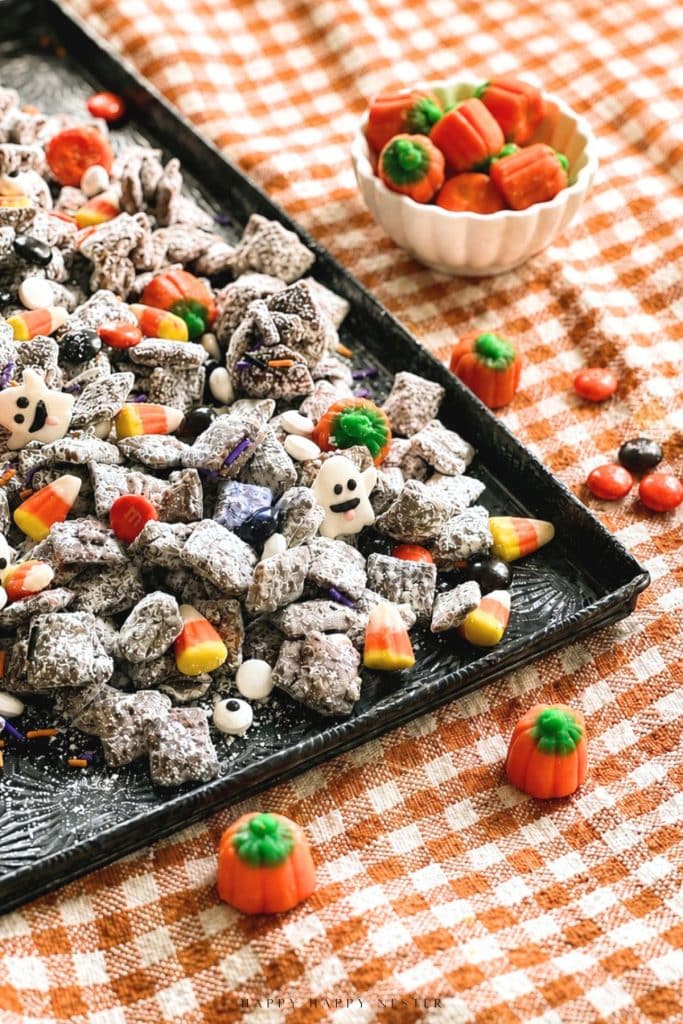 A tray of Halloween-themed puppy chow topped with candy corn, candy eyes, and ghost-shaped candies sits on a checkered tablecloth, offering the perfect halloween food inspiration with pumpkin-shaped candies scattered nearby.
