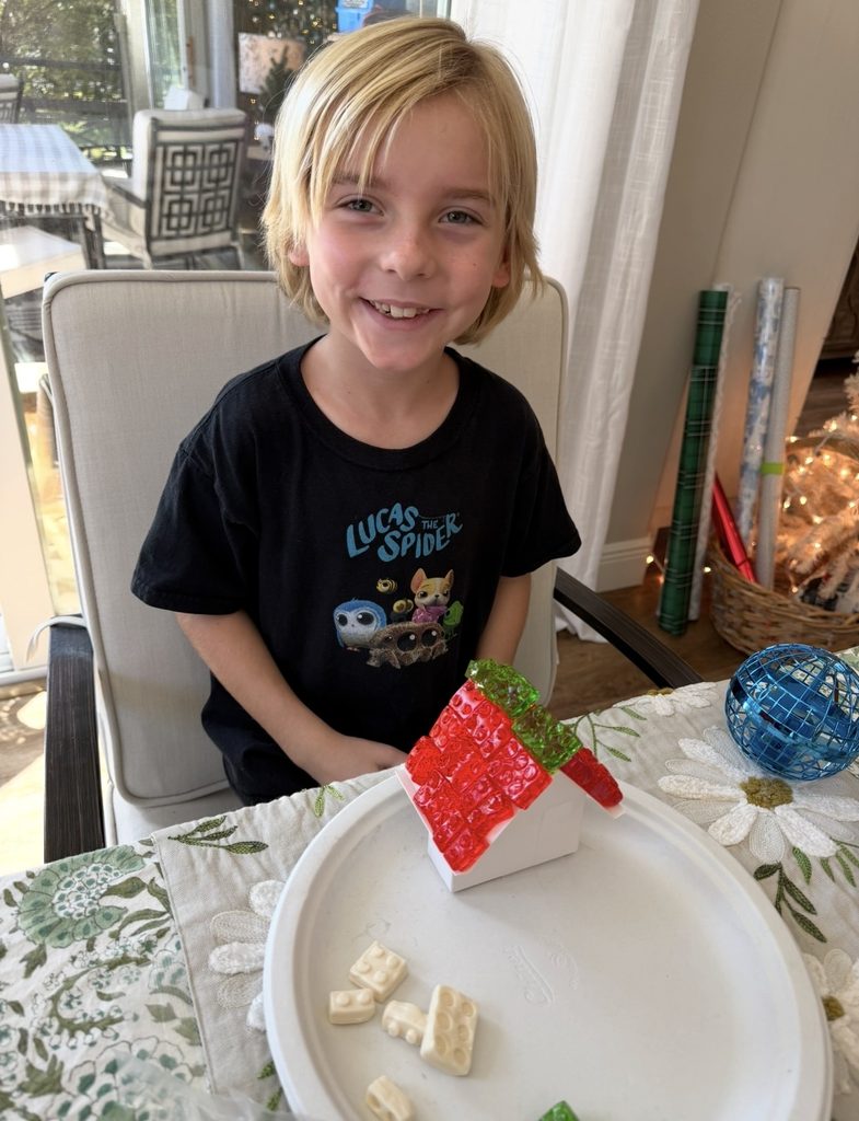 Smiling child with blond hair sits at a table, showing a small white house with a red and green roof, built from toy building blocks on a paper plate—perfect for a bit of Halloween food inspiration.