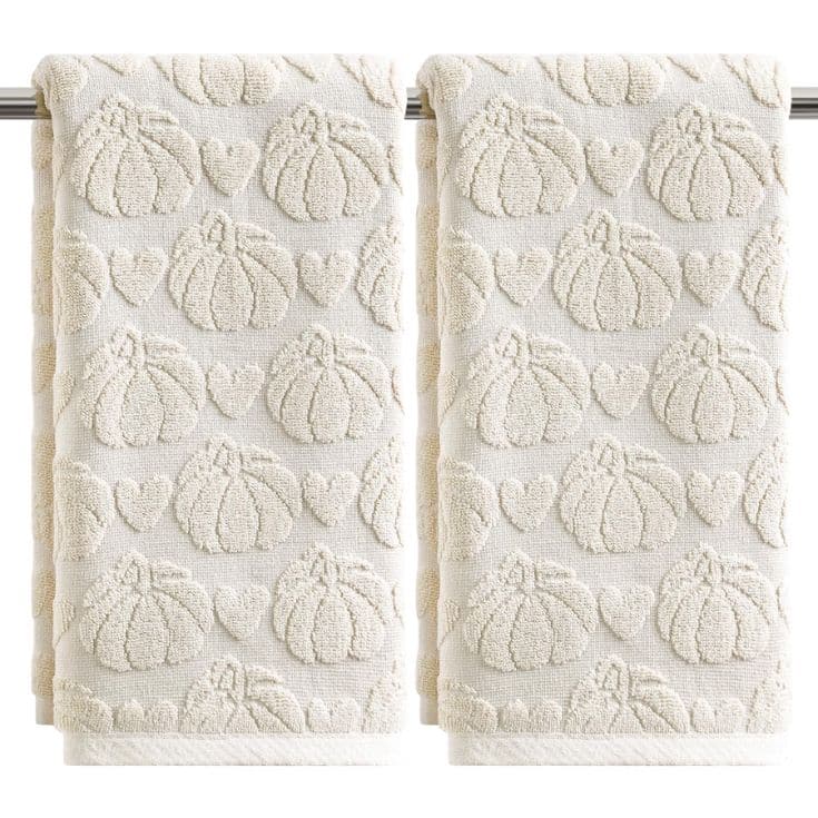 Two cream-colored towels hanging side by side on a rod, featuring a textured pattern of pumpkins and hearts throughout the fabric.