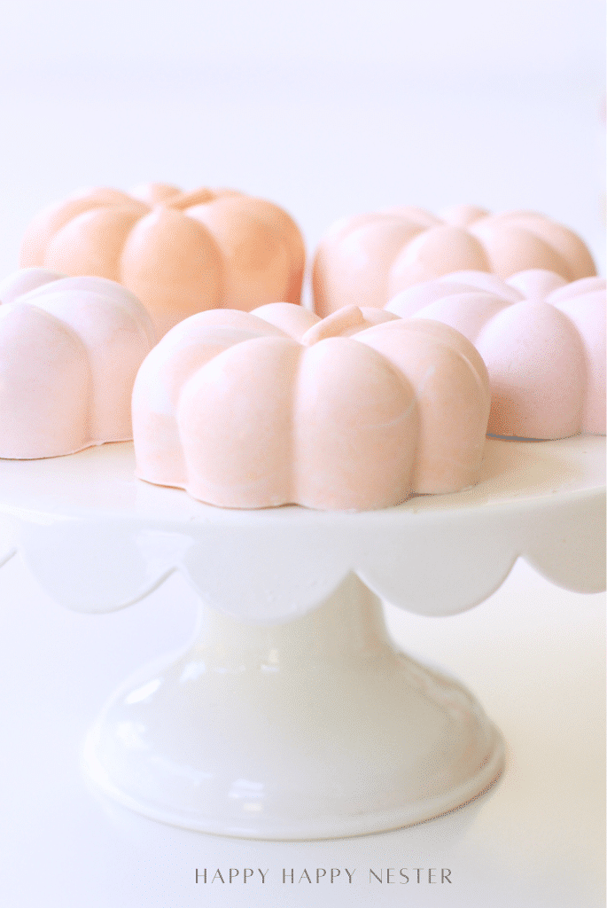 Four pastel pink pumpkin-shaped desserts sit on a white cake stand with scalloped edges against a light background, offering charming halloween food inspiration for your next festive gathering.