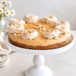 Easy no bake pumpkin cheesecake with graham cracker crust, cinnamon whipped cream, and small edible flowers on a white cake stand.