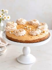 Easy no bake pumpkin cheesecake with graham cracker crust, cinnamon whipped cream, and small edible flowers on a white cake stand.
