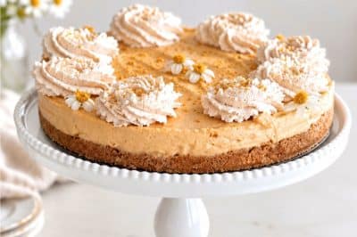 Easy no bake pumpkin cheesecake with graham cracker crust, cinnamon whipped cream, and small edible flowers on a white cake stand.