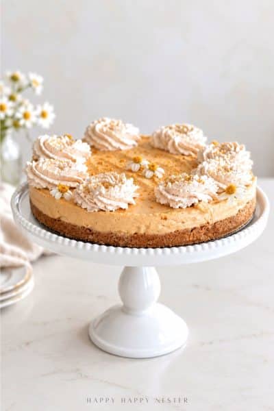 Easy no bake pumpkin cheesecake with graham cracker crust, cinnamon whipped cream, and small edible flowers on a white cake stand.