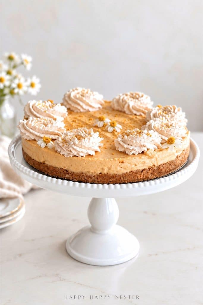 Easy no bake pumpkin cheesecake with graham cracker crust, cinnamon whipped cream, and small edible flowers on a white cake stand.