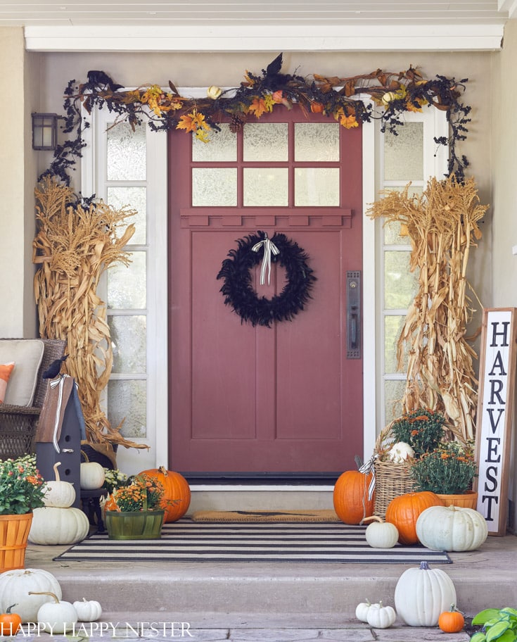 Fall front porch decorated with orange and white pumpkins, corn stalks, mums, garland, and a black wreath.