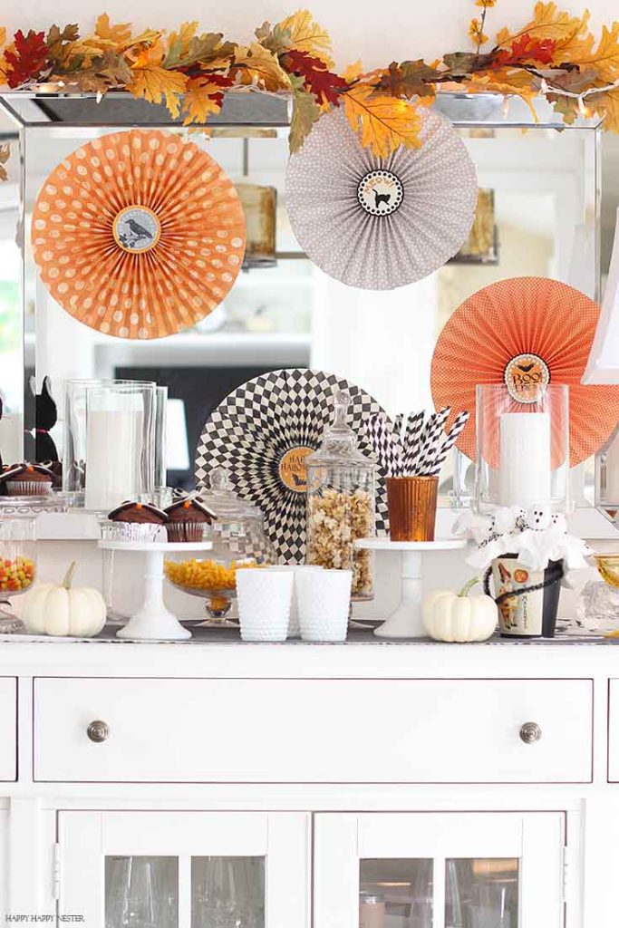 Halloween dessert station decorated with orange and black paper fans, white pumpkins, candy, cupcakes, and fall leaves.