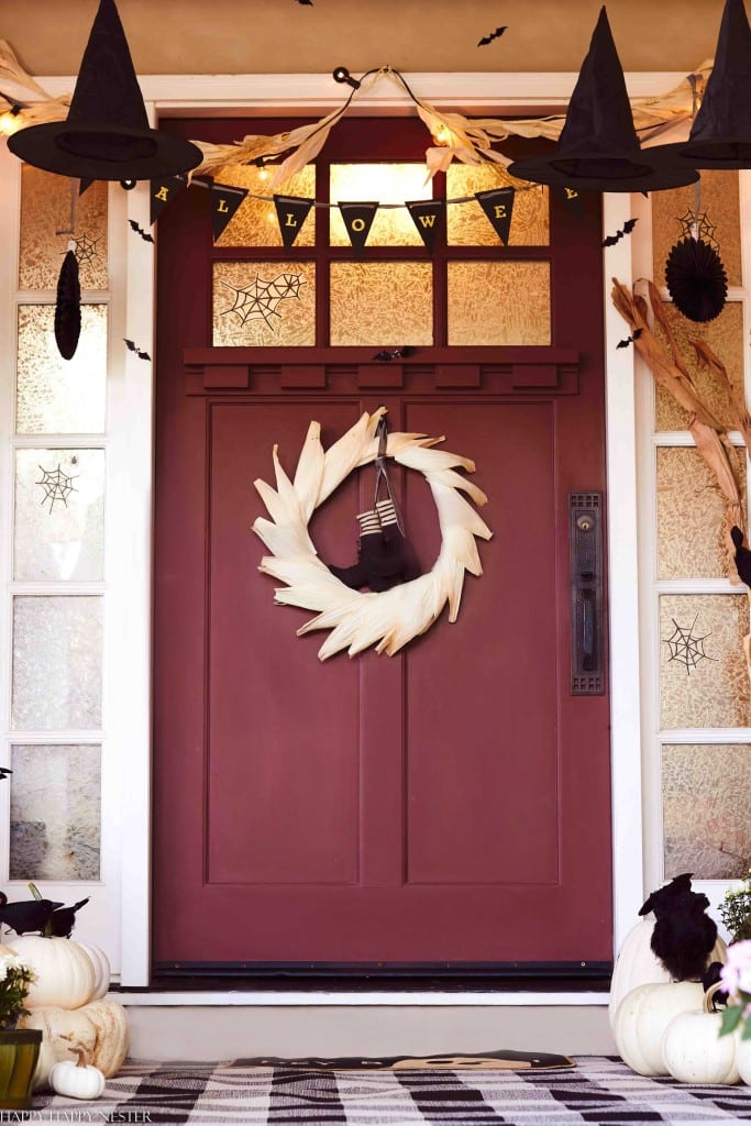 A maroon front door decorated for Halloween with a white wreath, black witch hats, bats, pumpkins, and a “HALLOWEEN” banner above showcases cute Halloween ideas. Pale pumpkins and a black-and-white checkered doormat are on the porch.