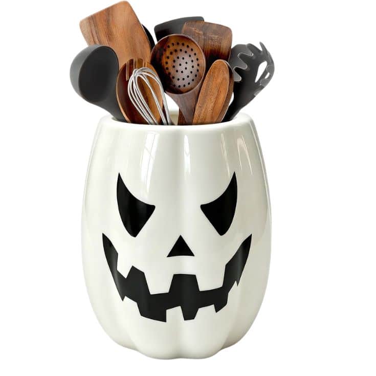 White ceramic container shaped like a pumpkin with a carved-face design, holding assorted wooden and metal kitchen utensils.
