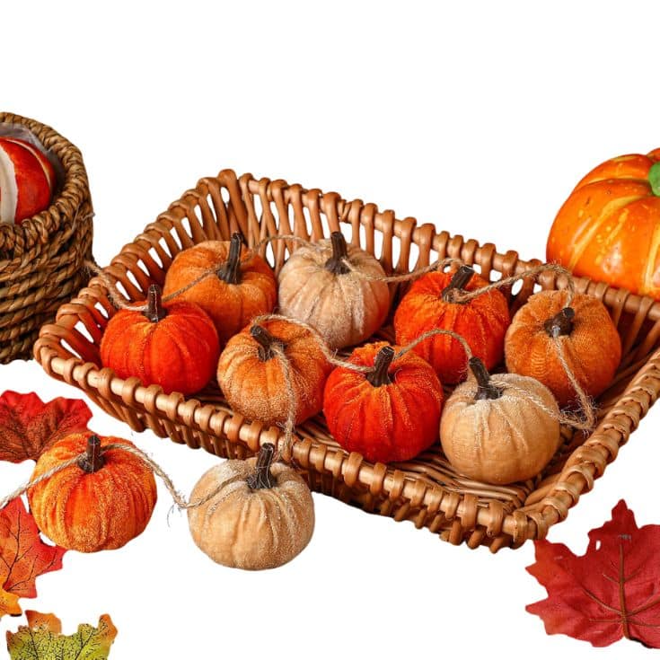 A wicker basket holds small fabric pumpkins in orange, beige, and brown. Autumn leaves and another basket are nearby, with a large orange pumpkin at the side, all set against a white background.