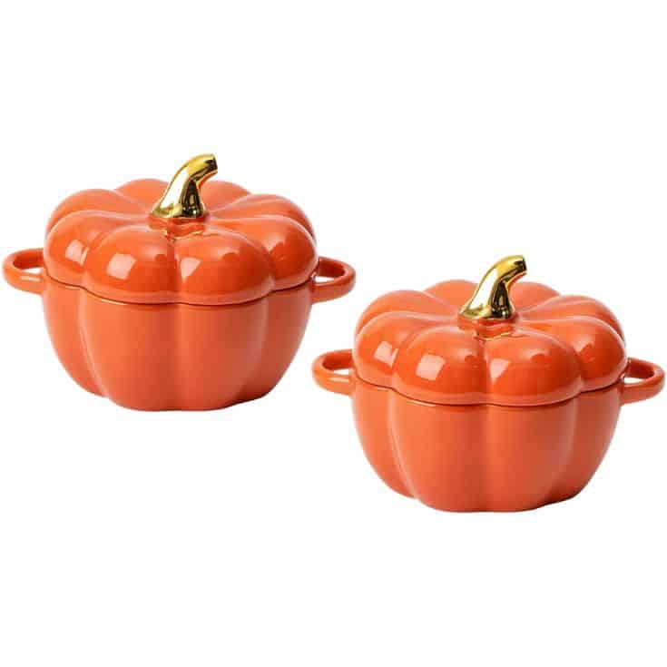 Two orange, pumpkin-shaped ceramic dishes with lids and gold-colored stems, each with two side handles, are shown against a white background.
