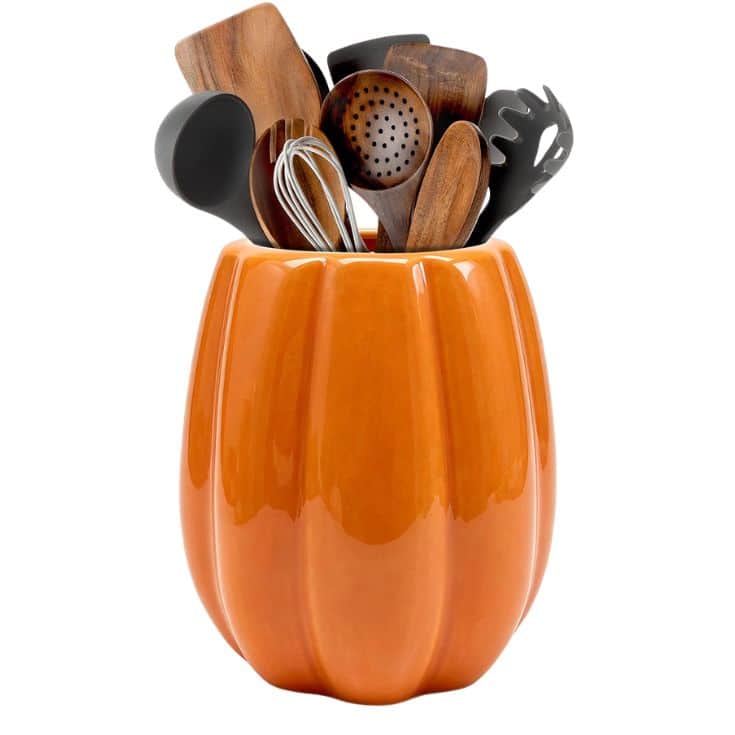 An orange, pumpkin-shaped ceramic container holds various kitchen utensils, including wooden spoons, a whisk, and black plastic spatulas, against a white background.