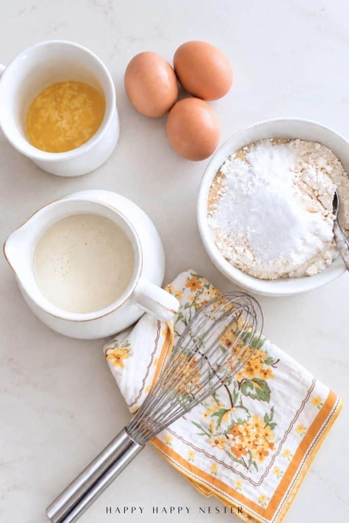 Ingredients for pancakes from scratch with eggs, buttermilk, melted butter, flour, sugar, and a whisk
