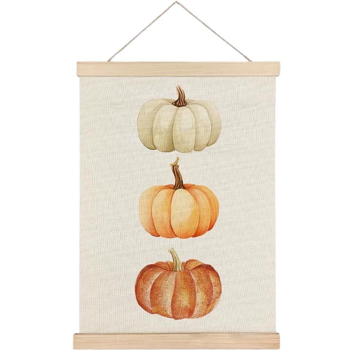 A hanging wall art features three pumpkins stacked vertically: a white pumpkin on top, an orange pumpkin in the middle, and a reddish-orange pumpkin at the bottom, all on a beige background with wooden hanger frames.