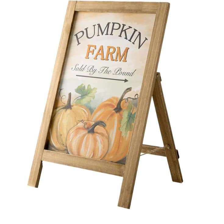Wooden A-frame sign with an illustration of pumpkins and text that reads, “Pumpkin Farm Sold By The Pound.”.