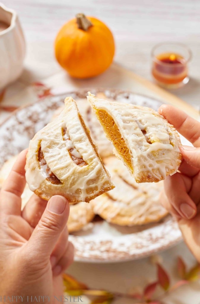 Hands holding a pumpkin hand pie broken in half, showing the orange filling. The hand pie is topped with white icing, offering perfect halloween food inspiration. A plate with more pies, a pumpkin, and a small jar of syrup are in the background.