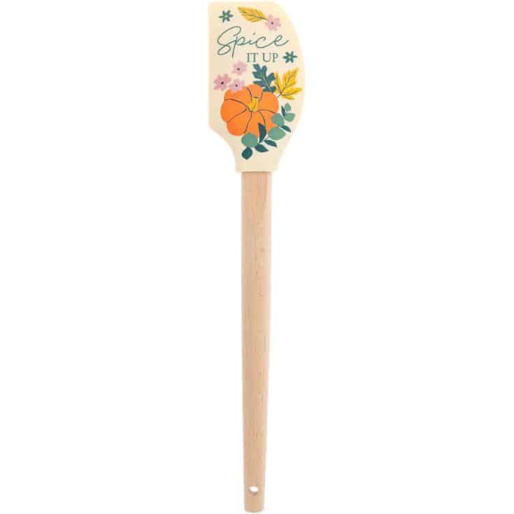 A spatula with a wooden handle and a decorated silicone head featuring a pumpkin, flowers, leaves, and the text “Spice it up.”.
