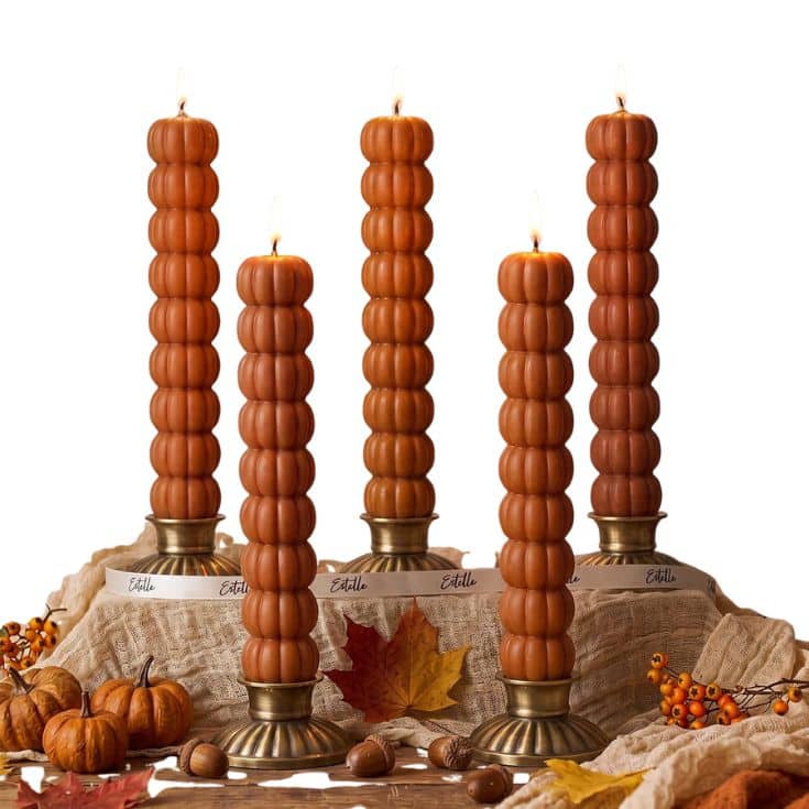 Five brown, pumpkin-shaped candles are lit and standing on brass holders, surrounded by autumn leaves, mini pumpkins, berries, and beige fabric on a white background.