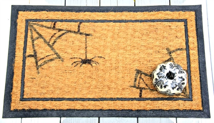 DIY Halloween doormat painted with black spider webs and spiders with a small decorative pumpkin.
