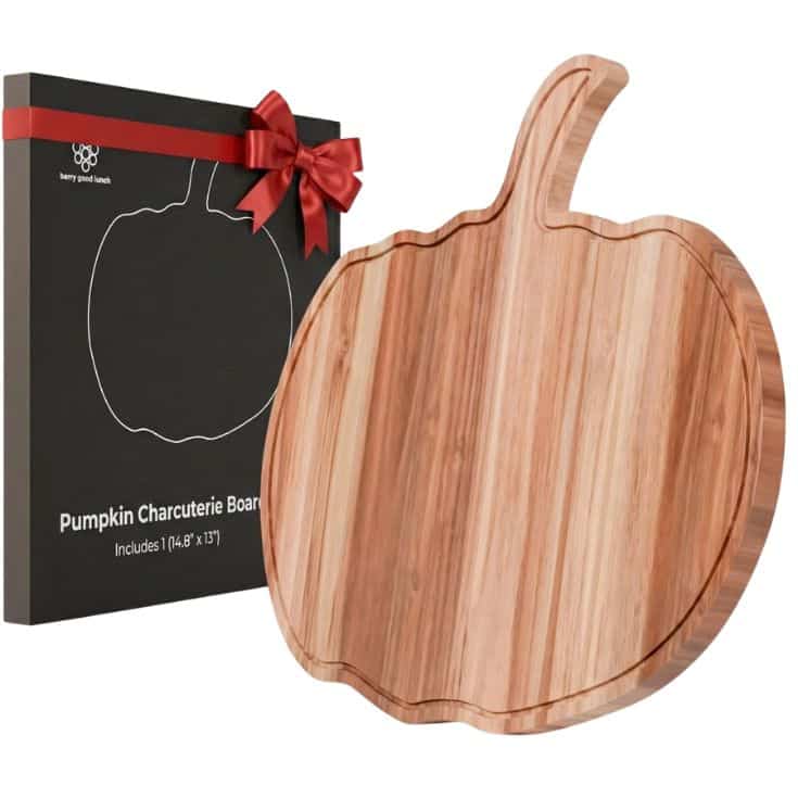 A wooden pumpkin-shaped charcuterie board is shown next to its black box packaging, which is tied with a red bow. The packaging displays an outline of a pumpkin and product description.