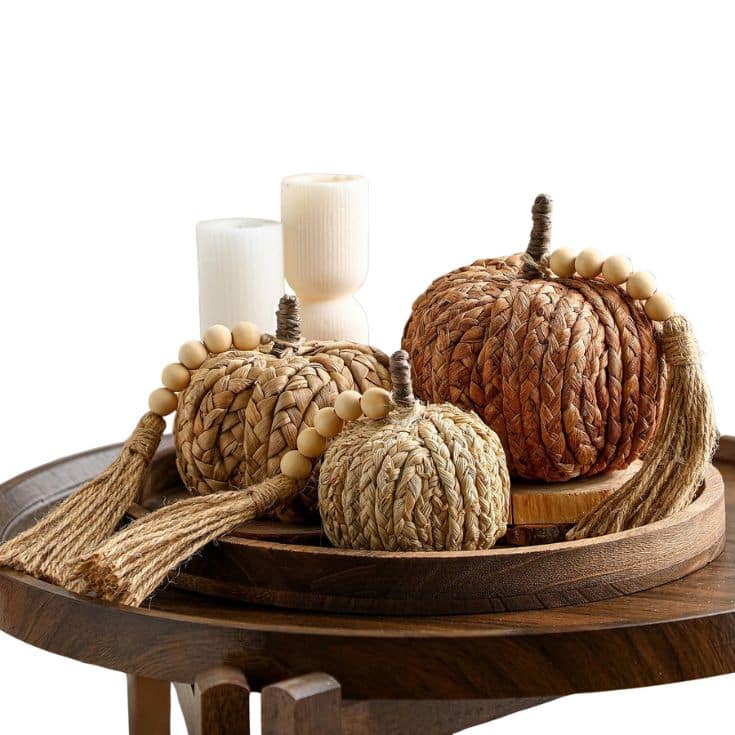 Three woven pumpkins with rope stems and beaded tassels sit on a wooden tray, alongside two white pillar candles, all placed on a wooden table against a white background.