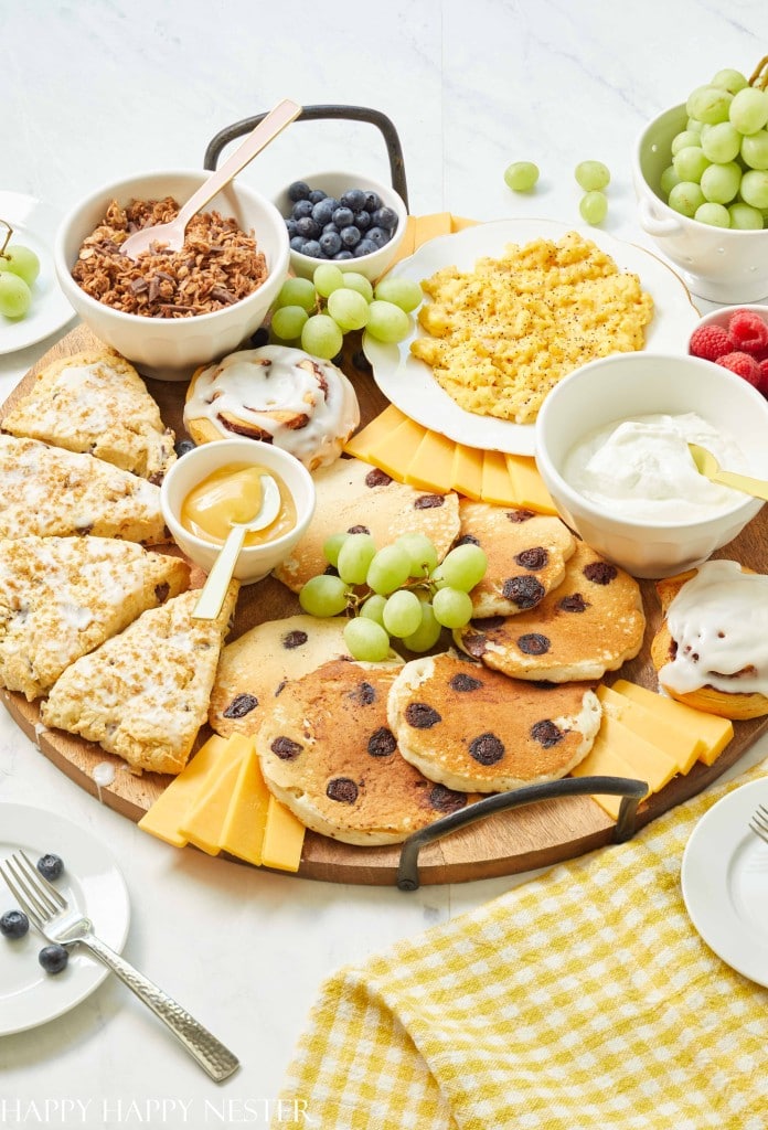 A large wooden serving board featuring holiday brunch ideas with blueberry pancakes, scones, scrambled eggs, sliced cheese, fresh fruit, granola, yogurt, and a cinnamon roll.