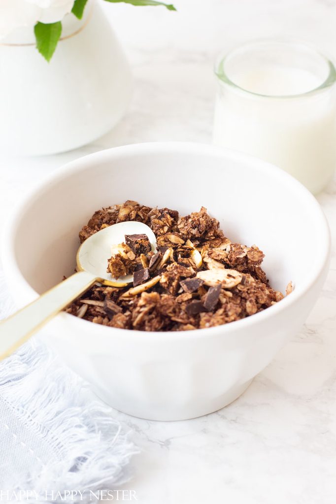 A white bowl of chocolate granola served for a holiday brunch, with a gold spoon on a marble surface next to a small jar of milk.