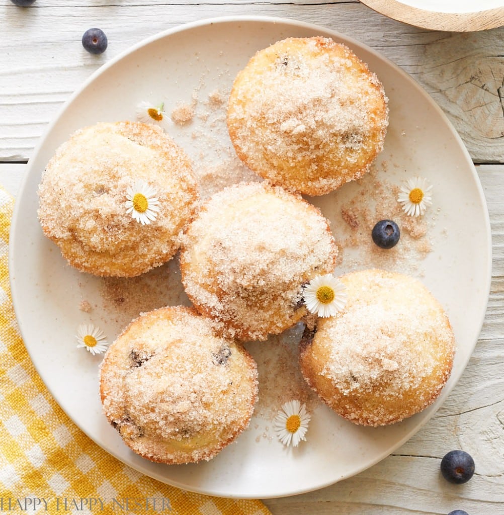 Five sugar-topped muffins for a holiday brunch, arranged on a plate and garnished with small white flowers and scattered blueberries.