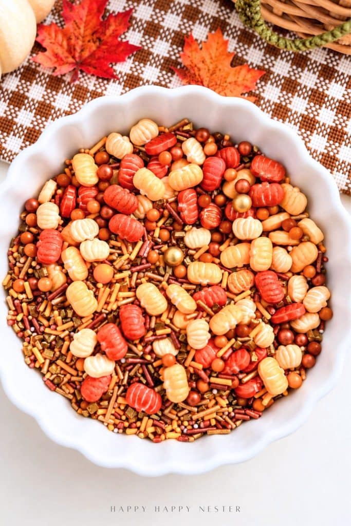 White scalloped bowl filled with orange and cream pumpkin-shaped candies, gold sugar pearls, and autumn-colored sprinkles for decorating fall sheet cakes.