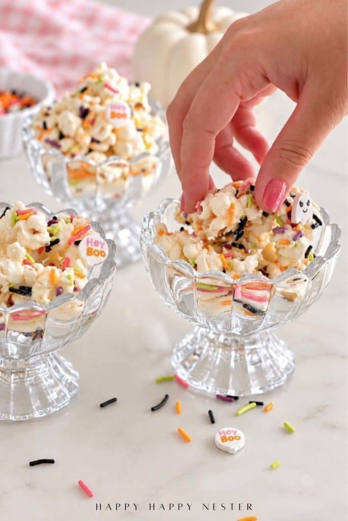 Hand reaching for coated popcorn with colorful sprinkles from a glass dessert bowl.