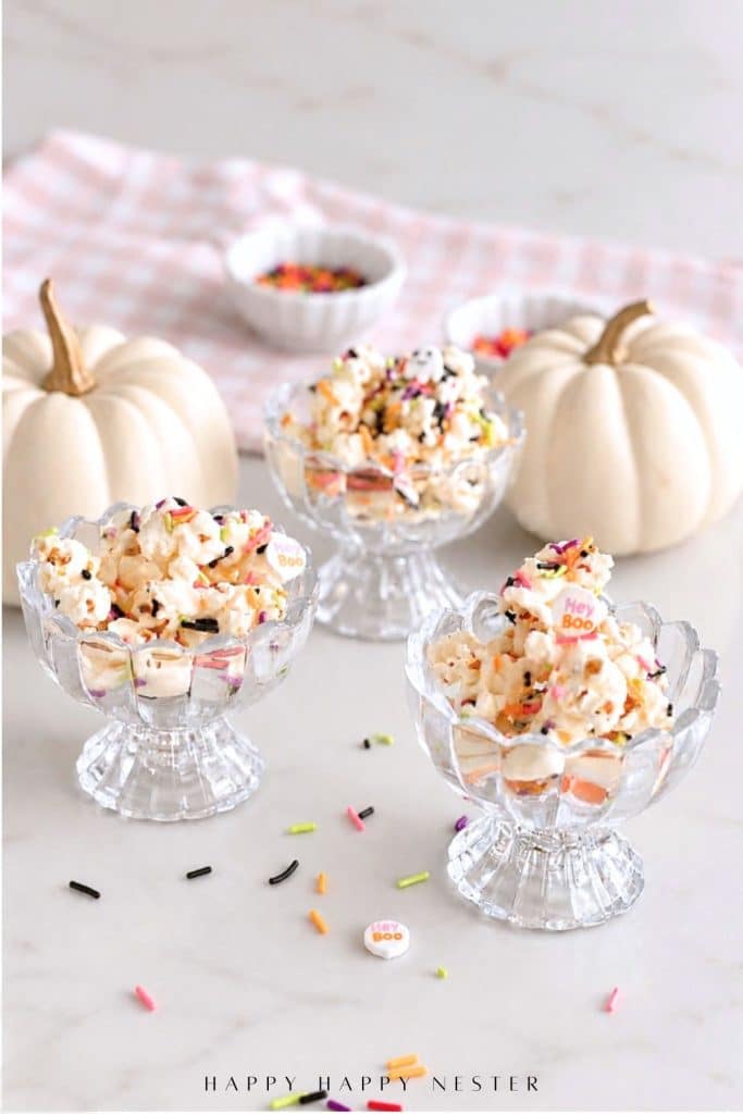 Three glass bowls filled with Halloween popcorn surrounded by white pumpkins and colorful sprinkles.